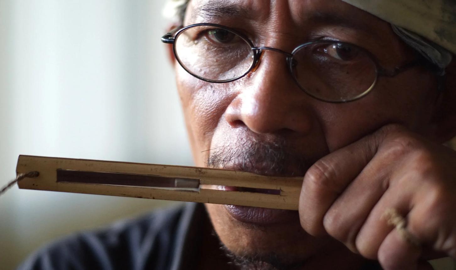 Foto close-up memperlihatkan bentuk fisik Kuriding yang dibuat dari pelepah enau (Arenga pinnata) atau bambu tipis. Alat musik ini termasuk kelompok jaw harp (harpa mulut). Bunyi dihasilkan dari getaran lidah (lamela) yang dipetik menggunakan jari, kemudian diperkuat oleh resonansi rongga mulut pemain sehingga menghasilkan nada-nada khas yang lembut dan ritmis.